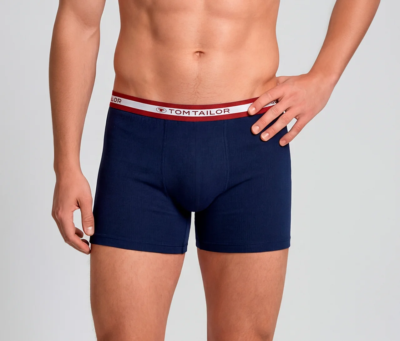 Mann trägt dunkelblaue Boxershorts mit weiß-rotem Tom Tailor Bund. Seine linke Hand liegt auf der Hüfte.