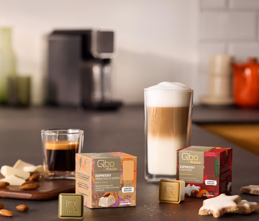Schwarze »Qbo ONE« Kapselmaschine im Hintergrund, davor ein Espresso, ein Latte Macchiato, zwei goldene Qbo Espresso Kapseln und zwei Packungen Qbo Espresso Kapseln „White Choc & Almond“ und „Zimtstern“ mit Zimtsternen, Mandeln und weißer Schokolade auf einer Küchenzeile.