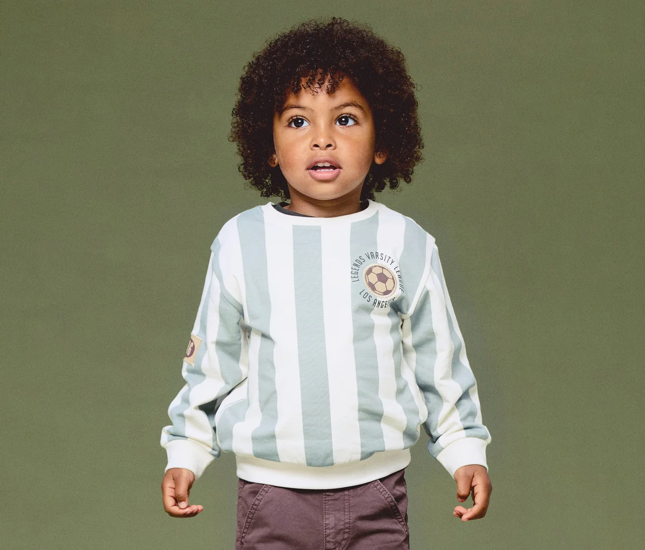 Ein kleiner Junge mit dunklen Locken trägt ein hellgrün-weiß gestreiftes Sweatshirt mit Fußball-Emblem und braune Hosen, blickt nach oben vor grünem Hintergrund.