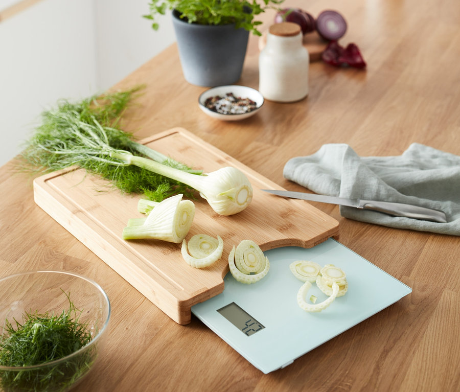 Auf einer Holzoberfläche liegt ein Schneidebrett mit herausnehmbarer Küchenwaage, auf der geschnittener Fenchel liegt.