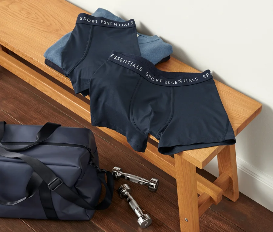 Zwei Sportslipboxer liegen auf einer Holzbank neben einer Sporttasche und Hanteln.