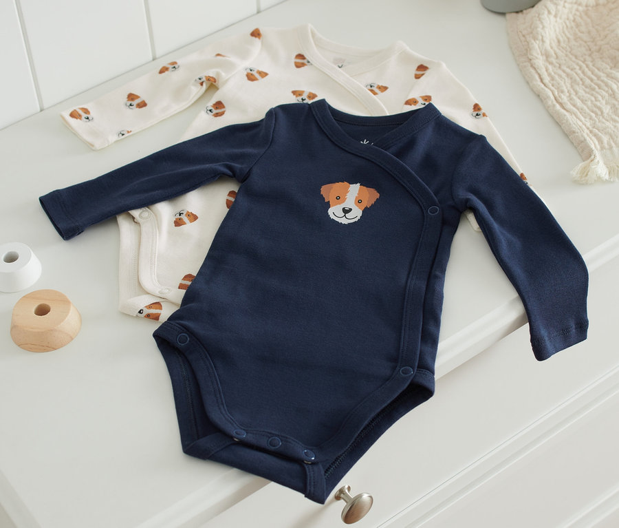 Zwei Babybodys liegen auf einer weißen Kommode. Ein Body ist dunkelblau mit einer Hundekopfstickerei, der andere ist cremefarben mit einem Hundeprint.