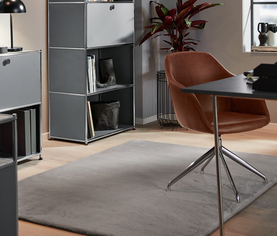 Im Raum stehen ein gepolsterter Drehstuhl aus Kunstleder, ein hohes Metallregal mit versetzbarem Klappenfach und eine LED-Metall-Tischleuchte. Auf dem Boden liegt ein Hochflorteppich.