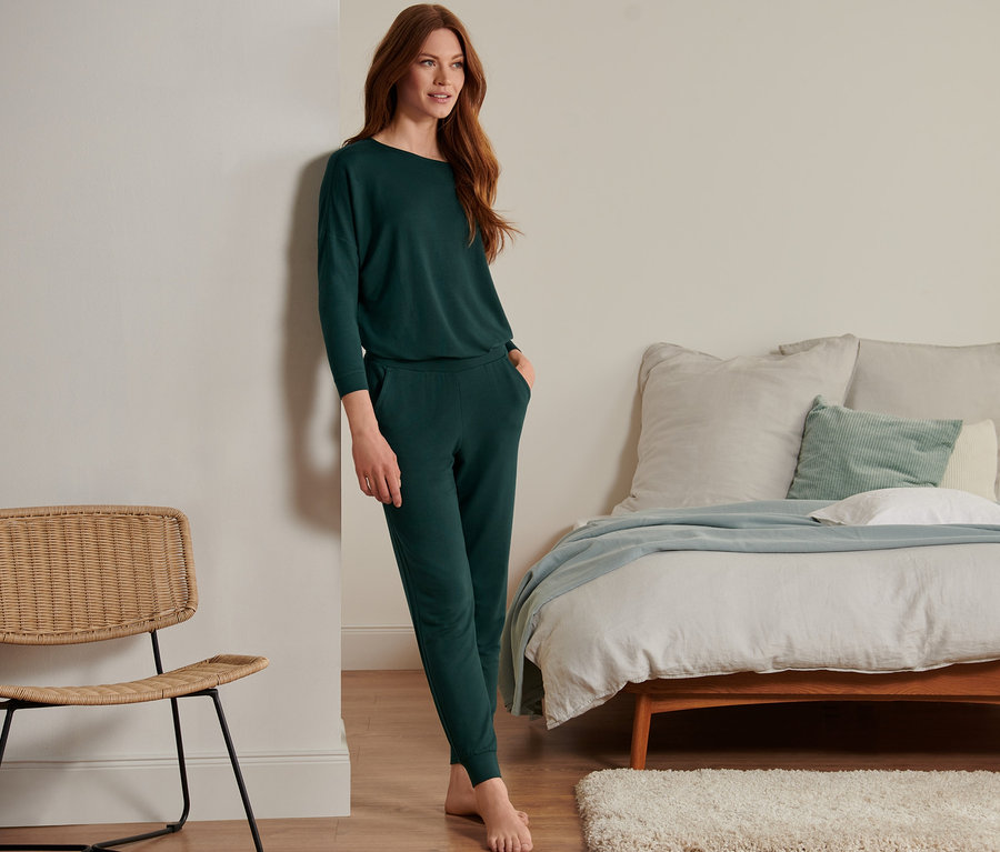 Eine Frau, die sich an eine Wand lehnt, trägt eine dunkelgrüne Homewear-Hose im Modal-Seide-Mix.