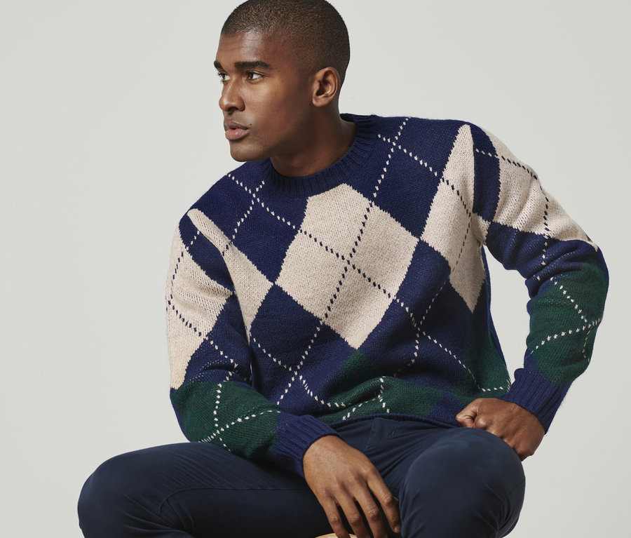 Mann sitzt auf einem Hocker und trägt einen Strickpullover.