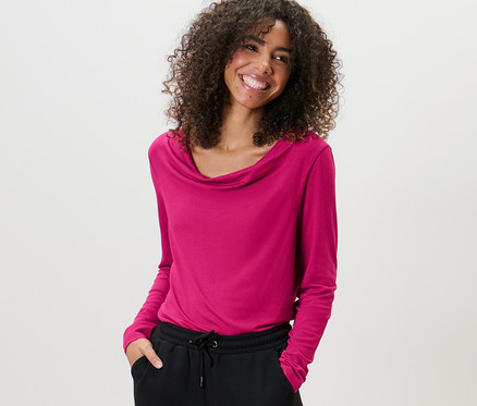 Frau mit lockigem Haar trägt ein Blusenshirt mit Wasserfallausschnitt und eine schwarze Hose.