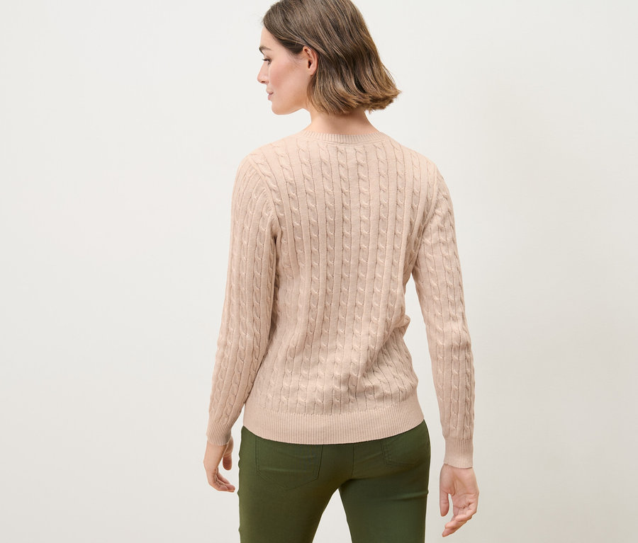 Frau mit braunen Haaren posiert mit dem Rücken zur Kamera und trägt einen cremefarbenen Zopfstrickpullover.