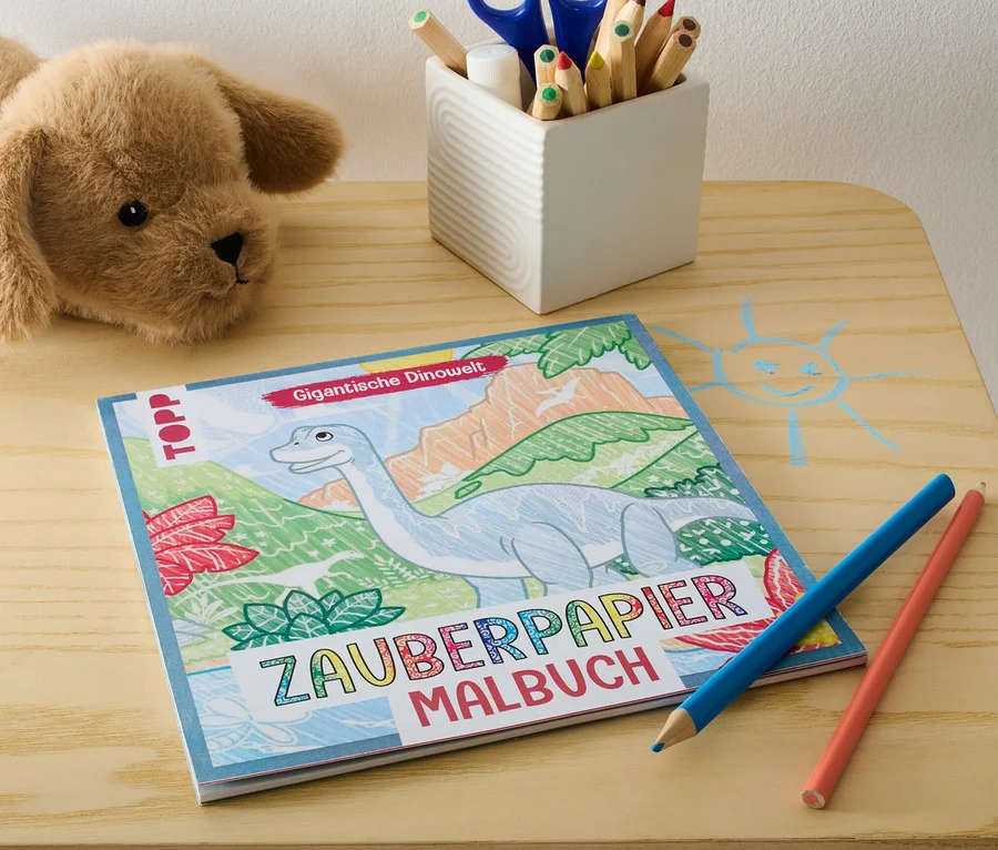 Auf einem Tisch liegen ein Plüschhund, das Buch "Zauberpapier Malbuch ‒ Gigantische Dinowelt", Buntstifte und ein Stiftehalter.