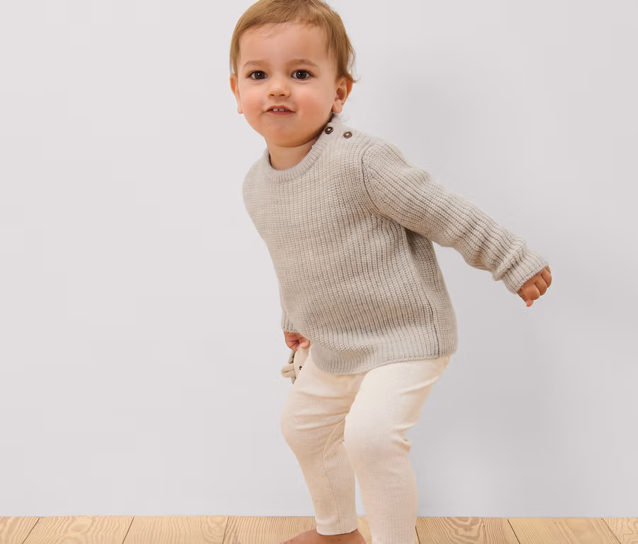 Kleinkind mit Kinder-Wollpullover und Leggings steht auf Holzboden.