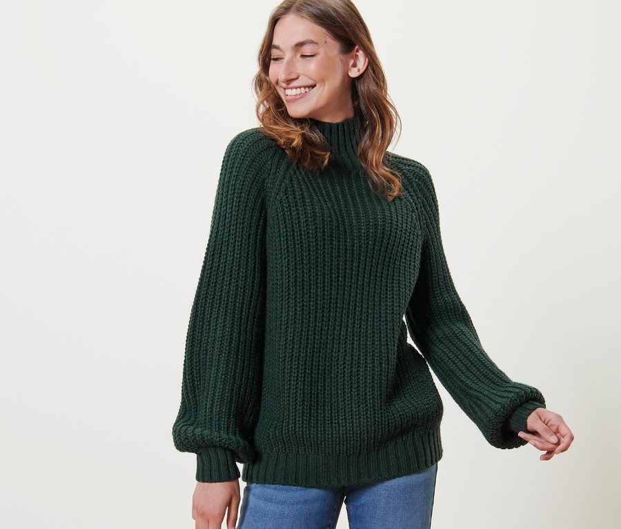 Frau mit braunen Haaren trägt einen dunkelgrünen Strickpullover mit Stehkragen und Jeans.