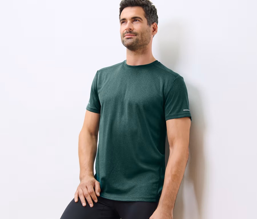 Mann lehnt an einer weißen Wand und trägt ein grün meliertes Funktionsshirt.