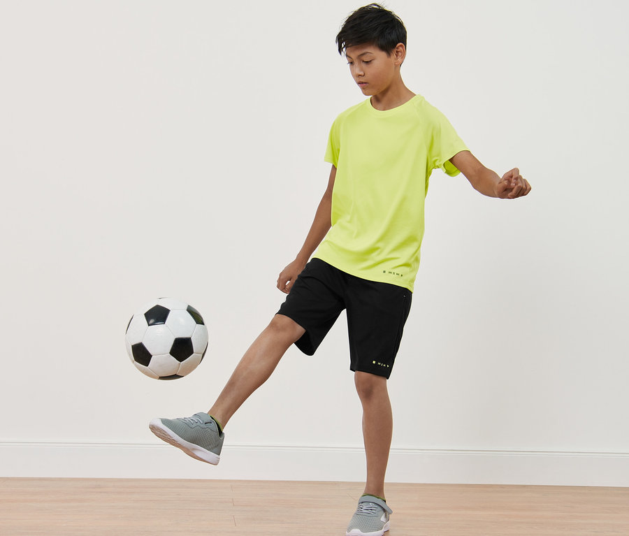 Ein Junge spielt mit einem Fußball. Er trägt schwarze Shorts und ein neongelbes Funktionsshirt. 2 Kinder-Funktionsshirts.