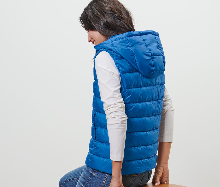 Frau sitzt auf einem Holzhocker und trägt eine blaue Steppweste mit Kapuze.