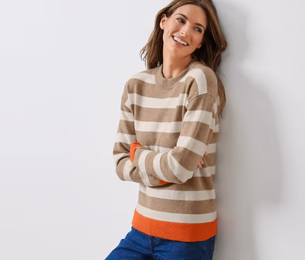 Frau lächelt an eine Wand gelehnt in einem gestreiften Strickpullover.