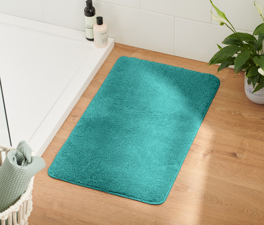 Türkise Badematte liegt auf Holzboden neben weißer Dusche und Weidenkorb mit Handtüchern.