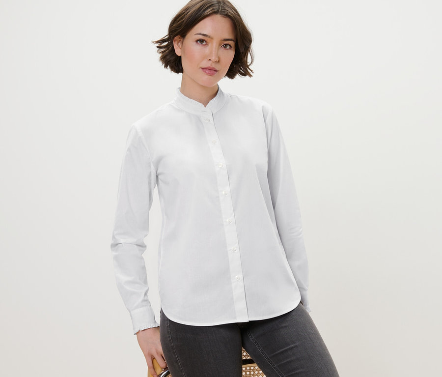 Eine Frau posiert auf einem Stuhl und trägt eine weiße Bluse mit Rüschendetail und graue Jeans.