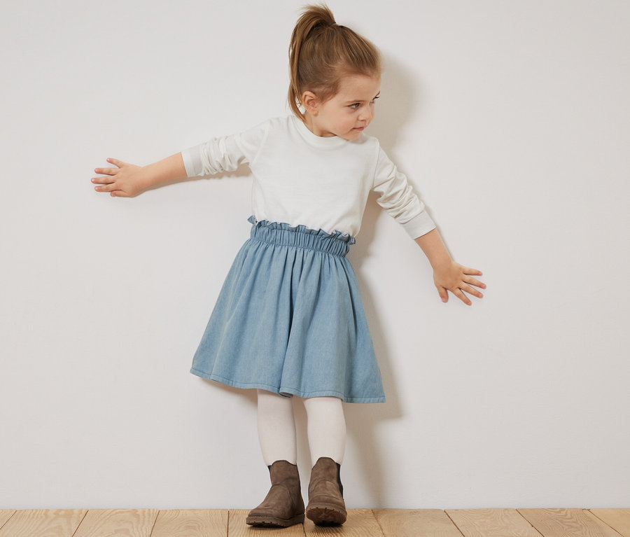 Mädchen posiert in Kinder-Denim-Wenderock, weißem Oberteil und braunen Stiefeln.