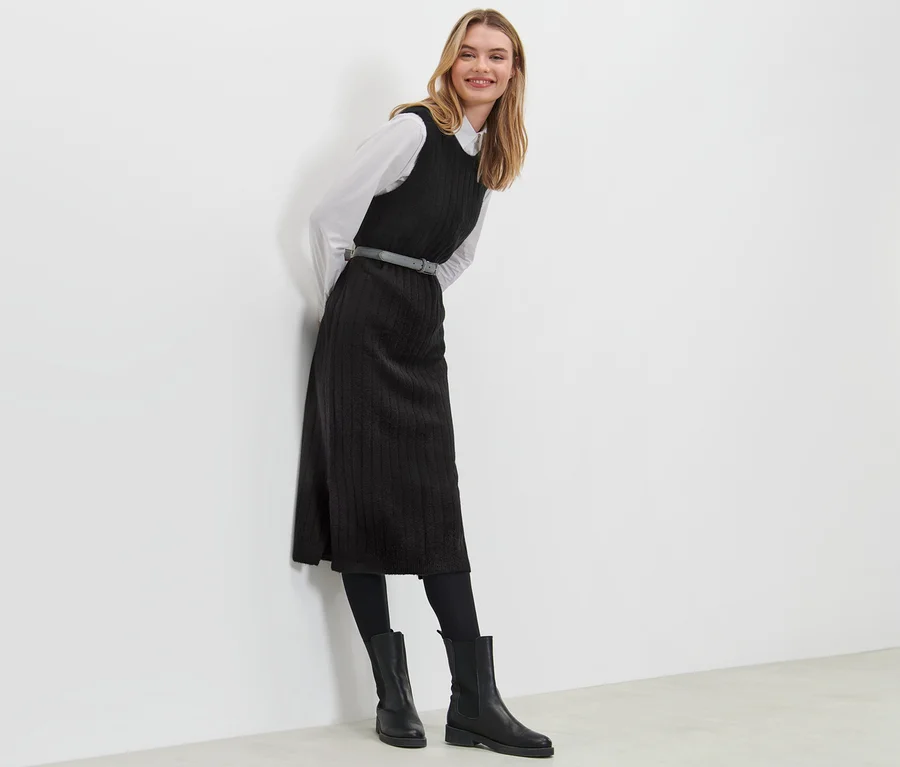 Frau, die an eine Wand gelehnt ist, trägt ein geripptes, schwarzes Strickkleid.