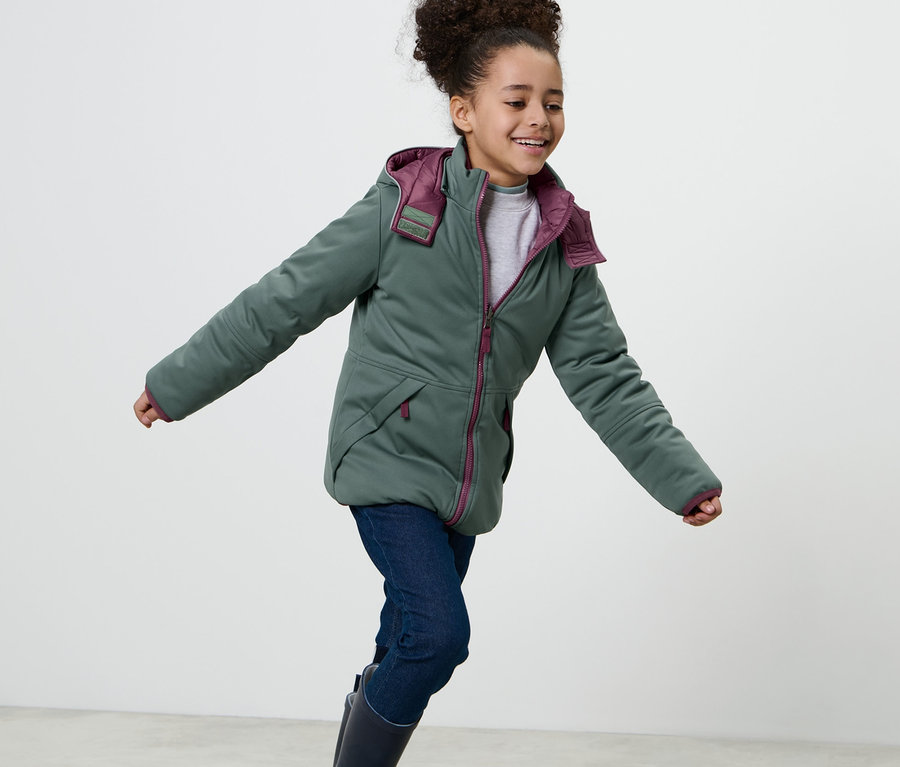 Lächelndes Mädchen in 2-in-1-Kinder-Softshell-und-Steppjacke mit Kapuze, Jeans und Gummistiefeln.