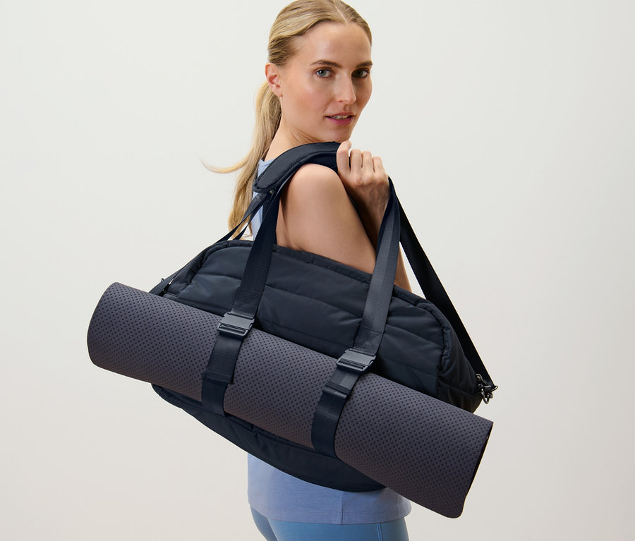 Blonde Frau trägt eine schwarze Sporttasche und eine Yogamatte über der Schulter.