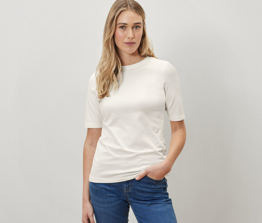 Frau posiert in einem cremefarbenen Halbarmshirt und blauen Jeans.
