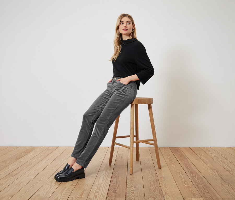 Blonde Frau sitzt auf einem Holzhocker, trägt ein schwarzes Langarmshirt, graue Cordhose und schwarze Slipper.