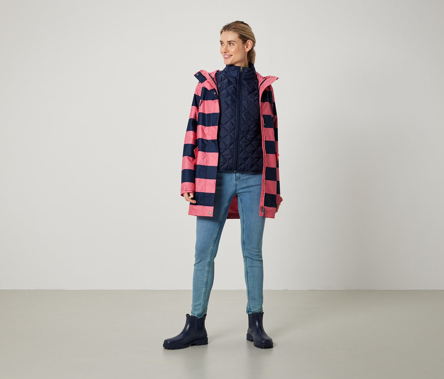 Eine Frau steht und trägt einen pink-marineblau gestreiften Mantel, eine marineblaue Weste, Jeans und blaue Stiefel.