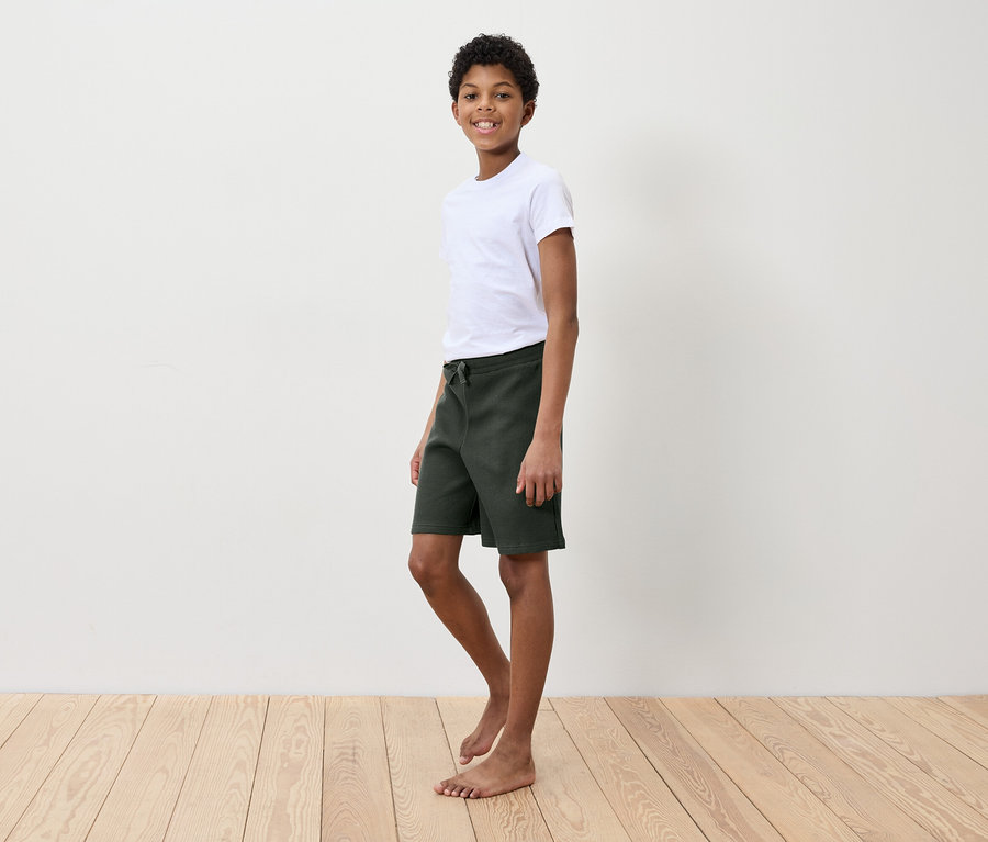 Ein Junge in einem weißen T-Shirt und grünen Shorts steht auf einem Holzboden vor einer weißen Wand.