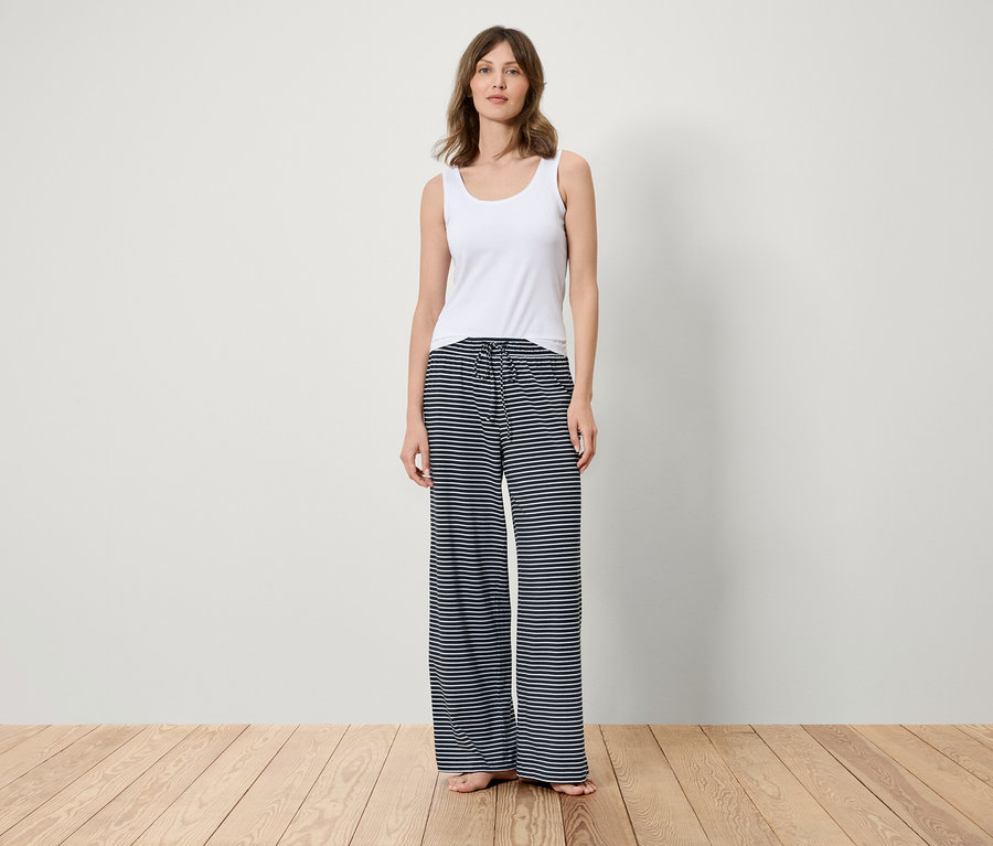 Eine Frau steht auf einem Holzboden und trägt ein weißes Tanktop und eine blau-weiß gestreifte Hose.