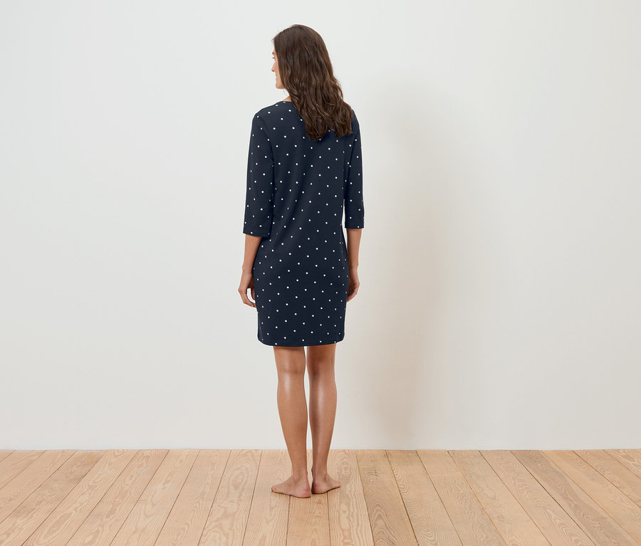 Eine Frau steht mit dem Rücken zur Kamera und trägt ein dunkelblaues Kleid mit weißen Punkten.