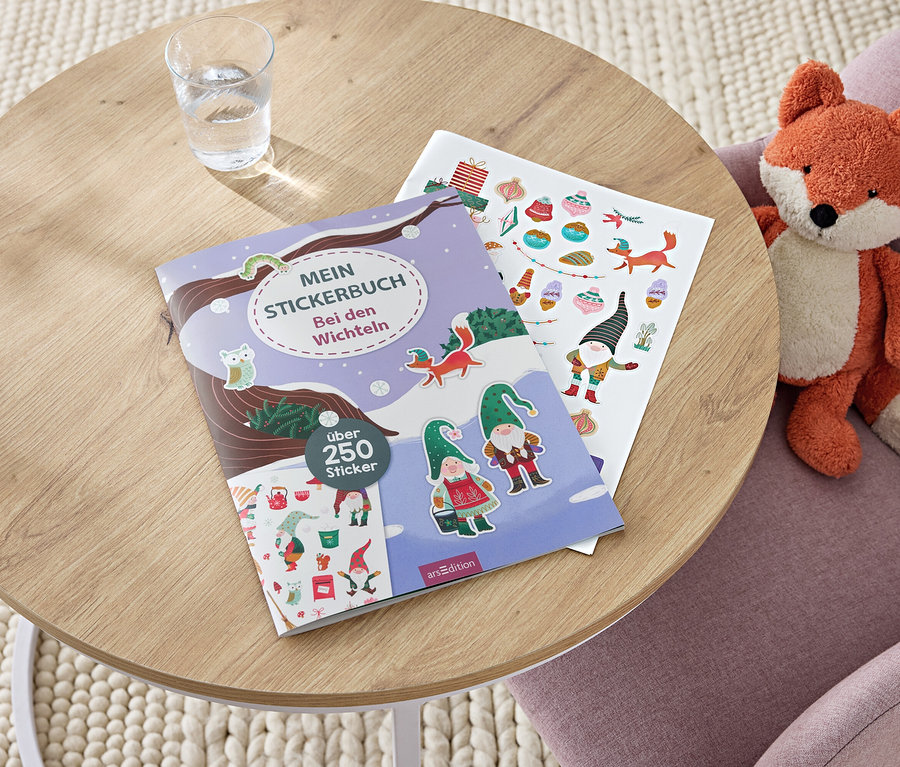 Auf einem Tisch liegen ein Aufkleberbuch, ein Bogen mit Aufklebern und ein Glas Wasser. Auf einem Stuhl sitzt ein Plüschfuchs.