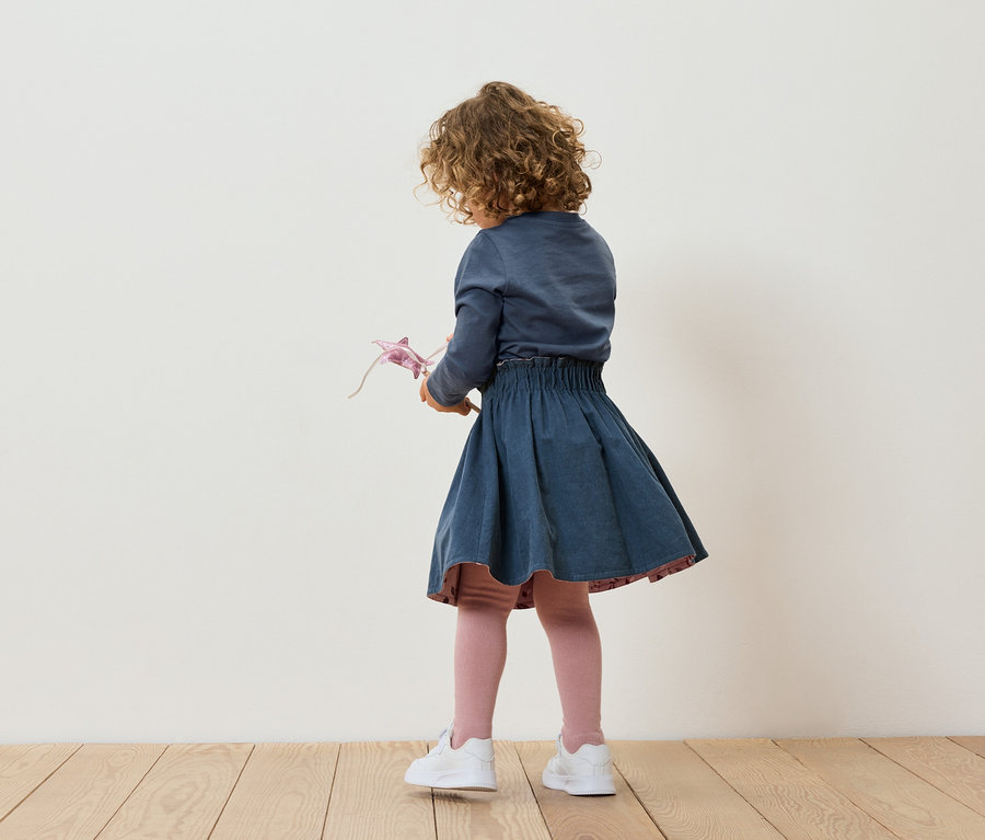 Mädchen mit lockigen Haaren steht mit dem Rücken zur Kamera, hält ein Spielzeug und trägt einen blauen Rock und weiße Turnschuhe.