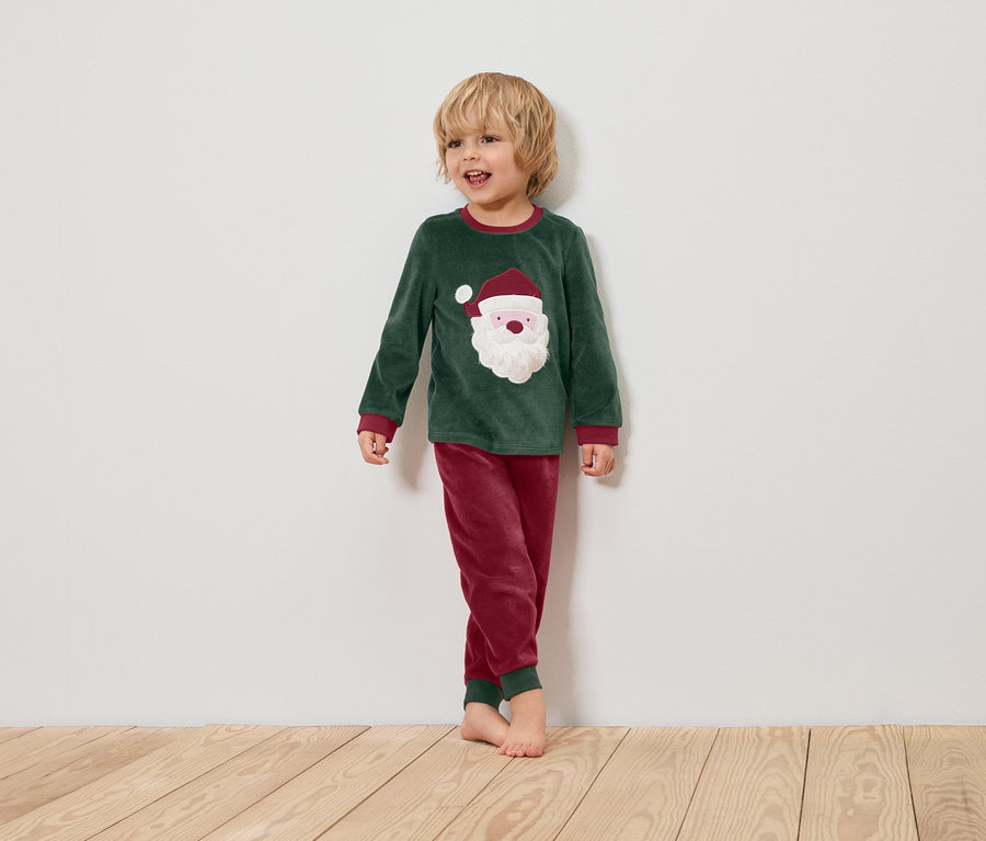 Ein Junge im grünen Weihnachtspyjama mit Santa-Claus-Motiv steht auf einem Holzboden vor einer weißen Wand.