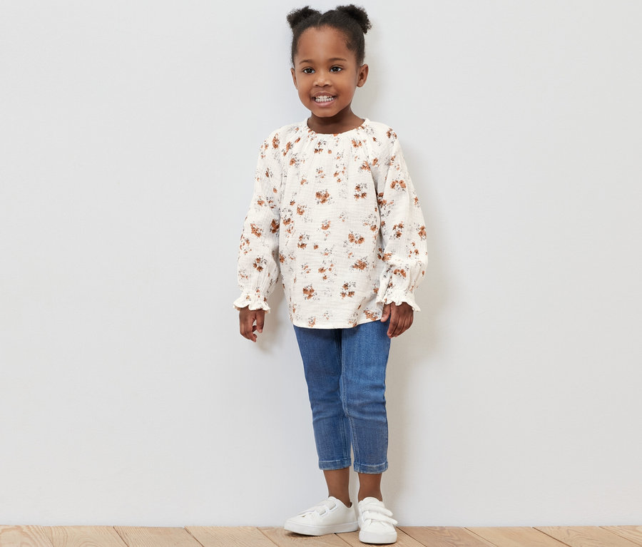 Lächelndes kleines Mädchen posiert im Studio und trägt eine Kinder-Musselin-Bluse, Jeans und Turnschuhe.