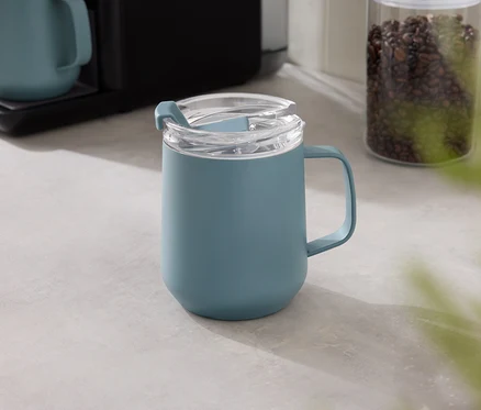 Eine blaue Tasse mit transparentem Deckel steht auf einer grauen Tischplatte, im Hintergrund eine Kaffeemaschine und ein Behälter mit Kaffeebohnen.