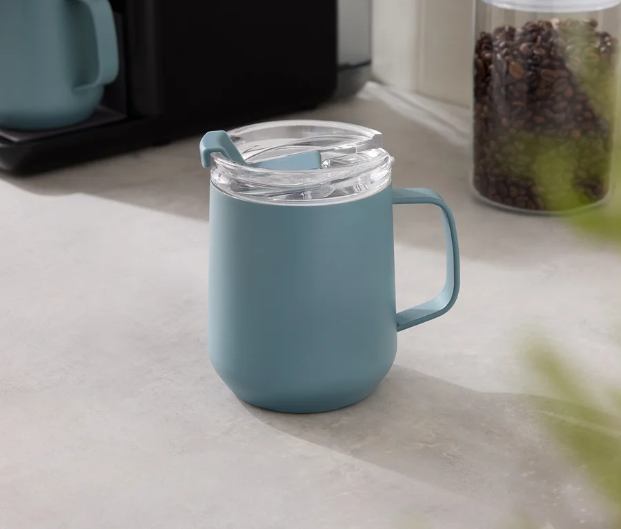 Eine blaue Tasse mit transparentem Deckel steht auf einer grauen Tischplatte, im Hintergrund eine Kaffeemaschine und ein Behälter mit Kaffeebohnen.