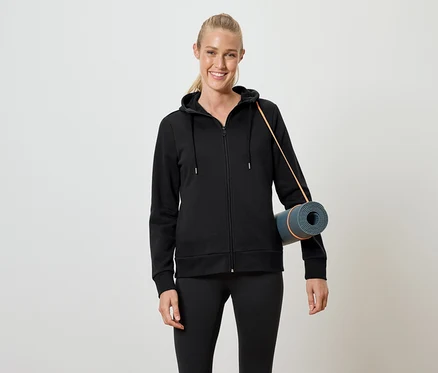 Frau in schwarzem Kapuzenpullover mit Reißverschluss und schwarzen Leggings, die eine zusammengerollte Yogamatte unter dem Arm hält.