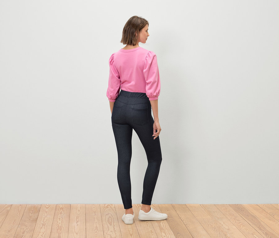 Eine Frau steht mit dem Rücken zum Betrachter und trägt ein rosa Oberteil mit Dreiviertelärmeln, dunkle Jeans und weiße Turnschuhe.