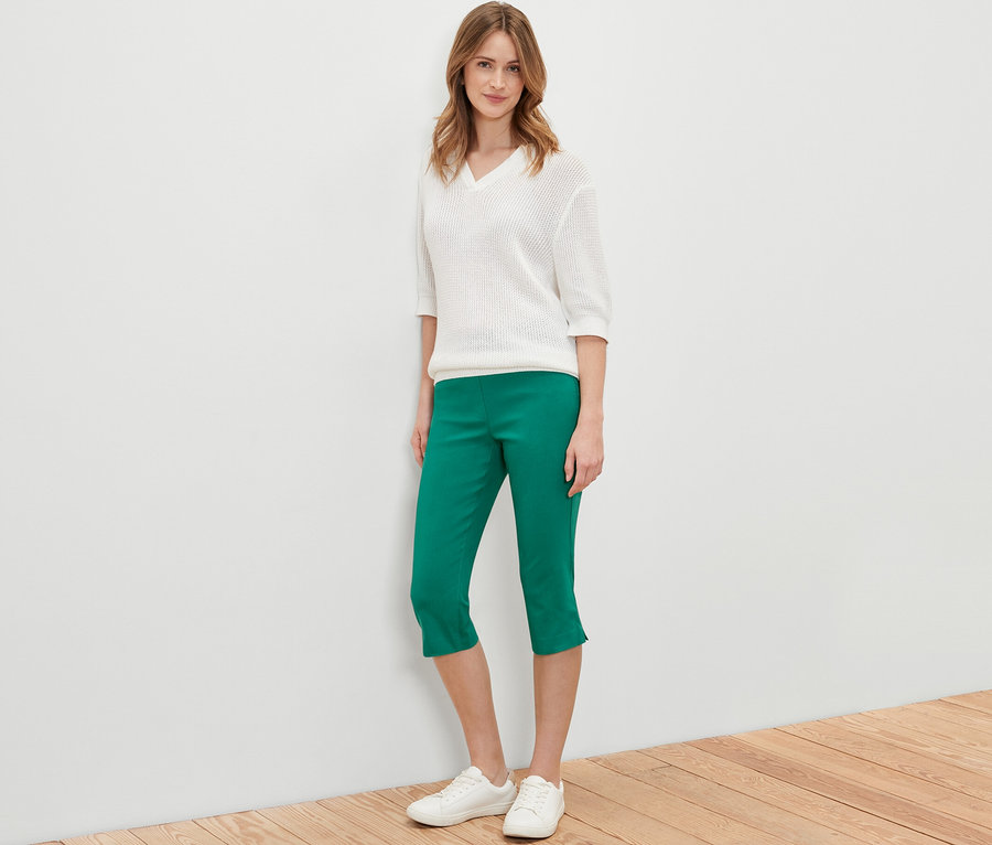 Frau posiert in grüner 3/4-Bengalin-Hose, weißem Pullover und weißen Schuhen.