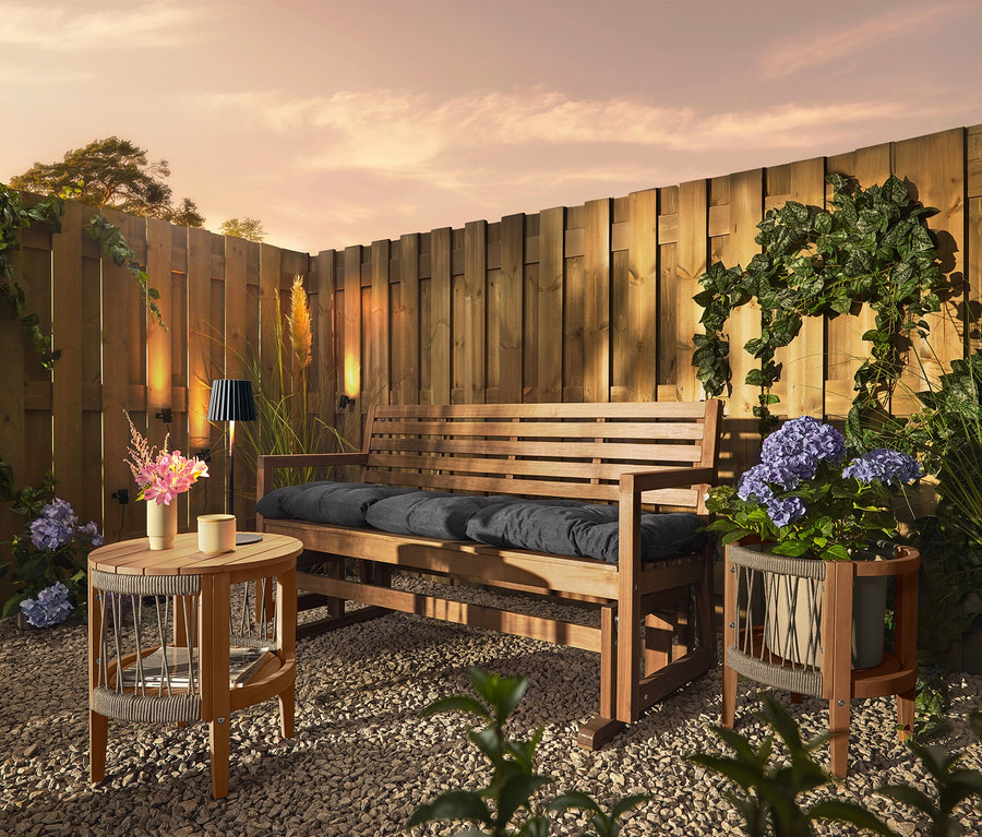 Eine Holzbank mit Kissen steht auf einem Kiesboden vor einem Holzzaun. An dem Zaun sind drei Solar-Dachrinnenleuchten befestigt.
