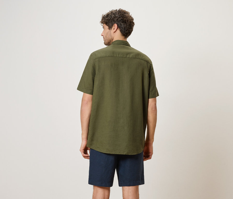 Ein Mann von hinten, trägt ein olivgrünes Hemd und blaue Shorts.