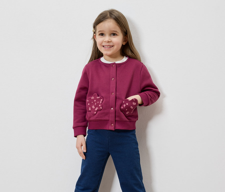 Lächelndes Mädchen mit braunen Haaren, trägt eine weinrote Strickjacke mit blumenförmigen Taschen und blaue Hosen vor einem weißen Hintergrund.