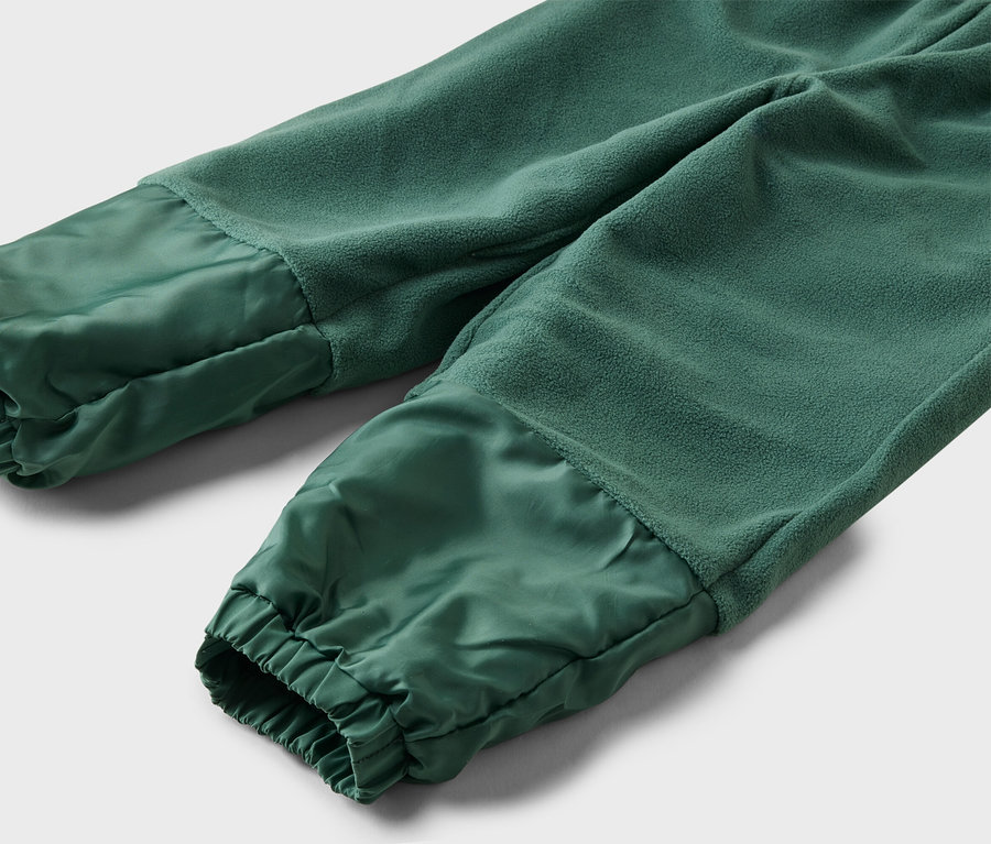 Nahaufnahme einer grünen Jogginghose mit Fleece-Oberteil und glänzendem, gerafftem Unterteil vor einem grauen Hintergrund.