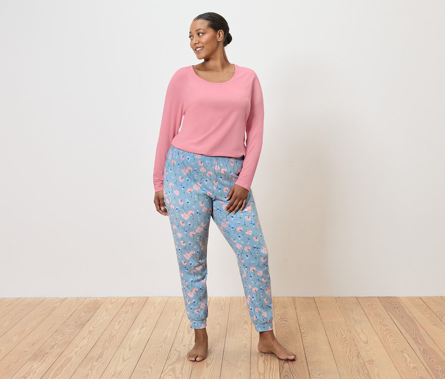 Frau steht barfuß, trägt einen pinkfarbenen Pullover und hellblaue Hosen mit Blumenmuster.