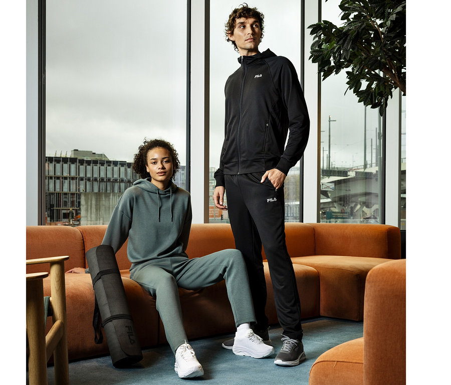 Ein Mann in einer schwarzen FILA Herren-Funktions-Sweatjacke steht neben einer Frau in grünem Hoodie und Jogginghose, die mit einer Yogamatte auf einer orangefarbenen Couch sitzt.