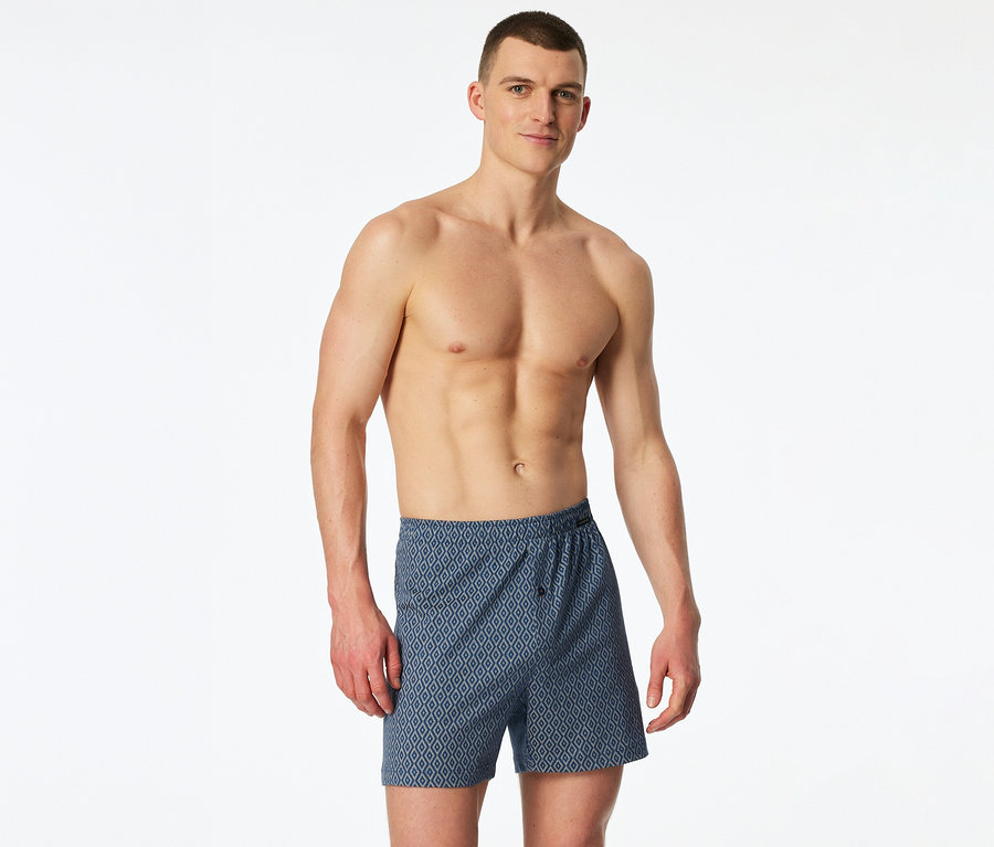Ein oberkörperfreier Mann trägt blaue SCHIESSER Jersey-Boxer mit Rautenmuster.