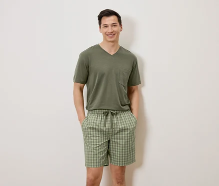 Ein Mann steht mit den Händen in den Taschen vor einer weißen Wand, er trägt ein olivgrünes T-Shirt und karierte Shorts.