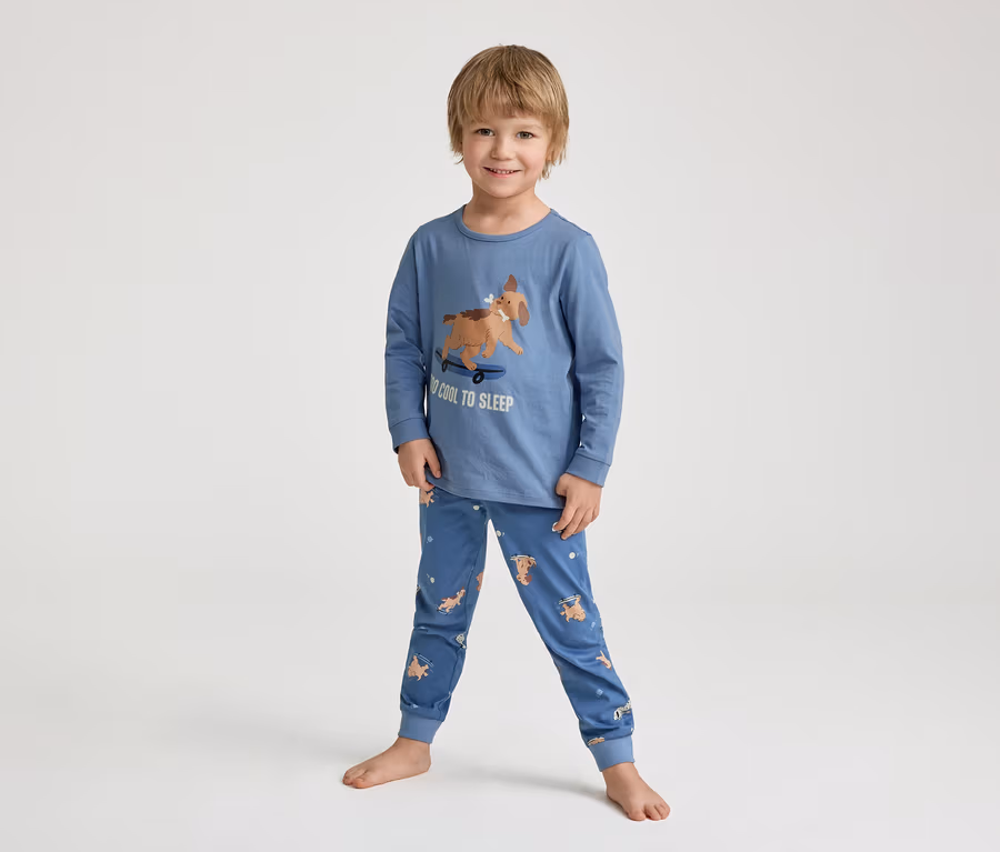 Ein lächelnder kleiner Junge steht barfuß in einem blauen Schlafanzug mit einem Hund auf einem Skateboard-Aufdruck und der Aufschrift "TOO COOL TO SLEEP".