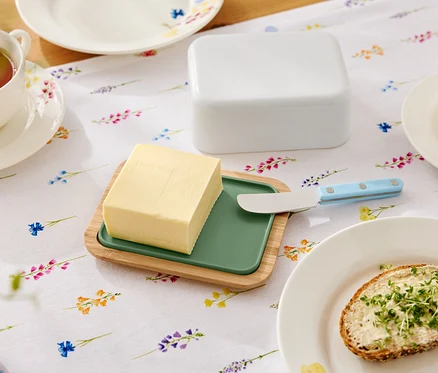 Auf einem blumigen Tisch liegt eine Butter auf einem hölzernen Teller mit grünem Einsatz, ein Buttermesser, eine Butterdose, eine Tasse Tee und ein Teller mit einem Butterbrot.