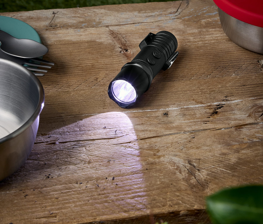 Eine schwarze Taschenlampe leuchtet auf einem Holztisch, neben einer Schüssel, Tellern und einem Behälter mit rotem Deckel.